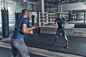 en el camino al éxito. apuesto joven africano vestido con ropa deportiva haciendo ejercicio usando pelotas mientras está parado frente al espejo en el gimnasio foto