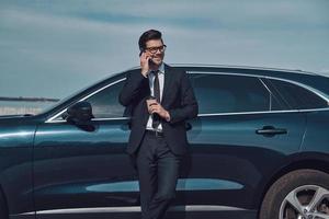 te estoy esperando a un apuesto joven hombre de negocios hablando por su teléfono inteligente y sonriendo mientras está parado cerca de su auto al aire libre foto