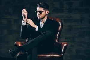 Everything must be perfect.Young handsome man in suit and sunglasses adjusting sleeve on his shirt while sitting in leather chair against dark grey background photo