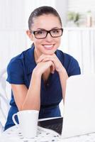 Smart and creative. Beautiful young woman holding hand on chin while sitting at the table with laptop and cup laying on it photo