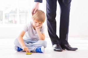 I will come back soon. Sad little boy leaning his face on hand while playing with toy car and looking down while his father in formalwear consoling him photo