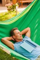 Relaxing in hammock. Happy mature man sleeping while lying in hammock with book laying on his torso photo