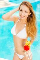 disfrutando de sus días de ocio de verano. vista superior de una atractiva joven en bikini blanco sosteniendo un cóctel y sonriendo mientras está de pie cerca de la piscina foto