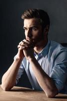 Just a moment to overthink everything. Side view of handsome young man keeping hands clasped and looking thoughtful while sitting against black background photo