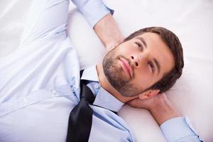 perdido en sus pensamientos. vista superior de un apuesto joven con camisa y corbata cogidos de la mano detrás de la cabeza y sonriendo mientras está acostado en la cama foto