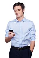 Always in touch. Handsome young man in formalwear holding mobile phone and smiling while standing isolated on white photo