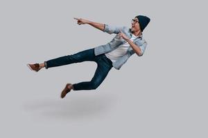 Pointing copy space in mid-air. Full length of handsome young man in jeans shirt gesturing and smiling while jumping against grey background photo