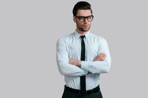 Confident in his style. Handsome young man in eyeglasses keeping arms crossed and looking at camera while standing against grey background photo
