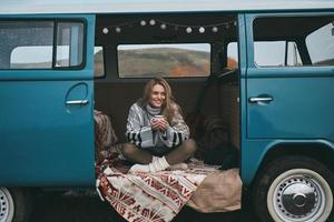 mejor viaje nunca. atractiva joven mujer sonriente sosteniendo una taza y mirando hacia otro lado mientras se sienta dentro de la mini furgoneta azul de estilo retro foto