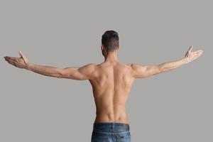 Man in perfect shape. Rear view of young muscular man keeping arms outstretched while standing isolated on grey background photo