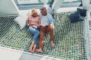 Beautiful mature couple examining smart phone and smiling while relaxing in big hammock at home together photo