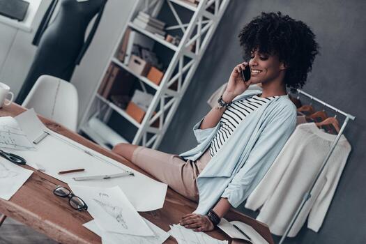 discutiendo las nuevas tendencias de la moda. bella joven africana hablando por teléfono inteligente y sonriendo mientras se sienta en el escritorio de su taller con ropa colgada en el fondo foto