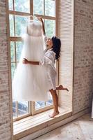 Special dress. Full length of beautiful young woman in silk bathrobe touching her wedding dress and smiling while standing near the window photo