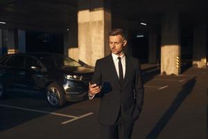 hombre de negocios maduro confiado mirando su teléfono móvil mientras está de pie en el estacionamiento foto