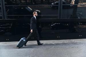 Business travel. Full length of young man in full suit pulling luggage while walking outdoors photo