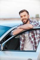 propietario de coche feliz. joven feliz apoyado en la puerta de su coche y sonriendo a la cámara mientras está de pie al aire libre foto