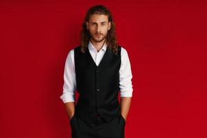 Charming young man in elegant clothing looking at camera and keeping hands in pockets while standing against red background photo