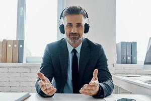 hombre de negocios confiado en auriculares sonriendo y gesticulando mientras está sentado en su lugar de trabajo foto