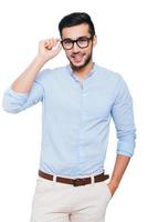 Charming handsome. Confident young Indian man adjusting his eyeglasses and smiling while standing against white background photo