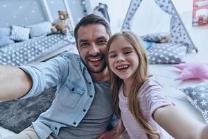 Family is the most important thing. Self portrait of young father and his little daughter smiling while sitting on the floor in bedroom with the tent in the background photo