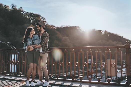 Great date. Full length of beautiful young couple embracing and looking at each other while standing on the bridge outdoors photo