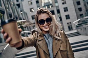 Attractive young woman carrying disposable cup and looking at camera while standing outdoors photo