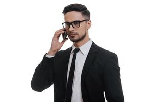 Important business talk. Serious young man in full suit talking on smart phone and looking away while standing against white background photo