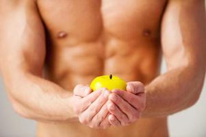 Living a healthy life. Close-up of young muscular man with perfect torso stretching out green apple while standing against white background photo