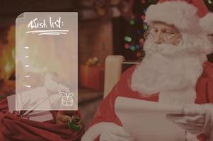 lista de deseos santa claus tradicional mirando su saco con regalos y sosteniendo un papel mientras se sienta en su silla con chimenea y árbol de navidad en el fondo foto