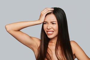 In pain. Frustrated young Asian woman making a face and keeping hand on head while standing against grey background photo