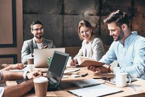 Brainstorming together. Group of young modern people in smart casual wear discussing business and smiling while sitting in the creative office photo