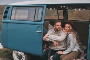 viaje romántico. hermosa pareja joven sonriendo y mirando hacia otro lado mientras se sienta en una mini furgoneta de estilo retro azul foto