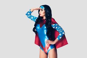 She can fight crime anywhere. Beautiful young woman in superhero costume looking away and keeping hand on forehead while standing against grey background photo