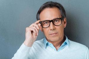 Confident mature man. Confident mature man adjusting his eyewear and looking at camera while standing against grey background photo