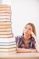 Tired of studying. Depressed teenage girl holding hand on chin and looking at the top of the big stack of books laying on the desk photo