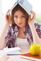 Feeling tired of studying. Frustrated teenage girl carrying book on head and looking at camera while sitting at the desk with books laying on it photo