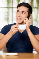 descanso. alegre joven bebiendo café en el restaurante y hablando por teléfono móvil foto