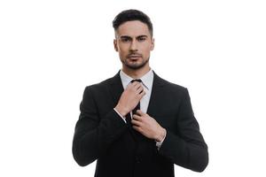 Always perfect. Handsome young man in full suit adjusting his necktie and looking at camera while standing against white background photo