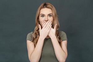 Trying to stop gossiping. Surprised young woman in casual wear covering mouth with hands and looking at camera while standing against grey background photo