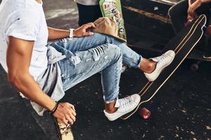 Skating is his life. Close-up of young modern man in casual wear keeping feet on his skateboard while spending time with friends outdoors photo