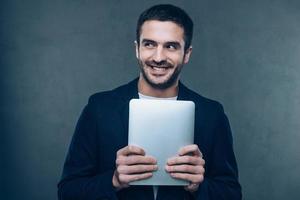 mi precioso. un joven alegre sosteniendo su tableta digital y sonriendo mientras se enfrenta a un fondo gris foto