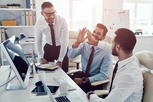 High five for success. Group of young modern men in formalwear smiling while working in the office photo