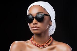 combinando moda y tradiciones. hermosa mujer africana con un pañuelo en la cabeza y gafas de sol mientras está de pie contra el fondo negro foto
