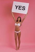 Full length of attractive young woman holding a poster and smiling while standing on tiptoe against pink background photo