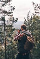 comprobando la ruta. joven moderno con mochila mirando hacia otro lado a través de binoculares mientras está de pie en el bosque foto