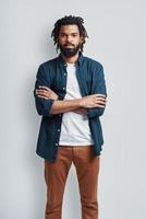 Thoughtful young African man in casual wear looking at camera and keeping arms crossed while standing against grey background photo
