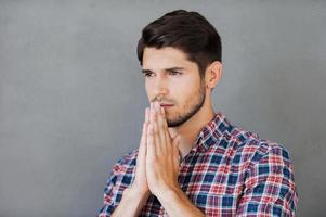 Begging for good. Thoughtful young man holding hands clasped near face and looking away while standing against grey background photo