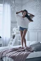 Feeling free to do everything. Full length of young woman keeping hands in hair with closed eyes while standing on the bed at home photo