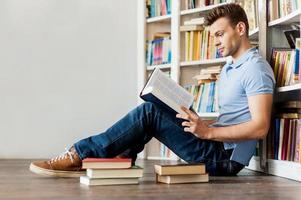 Young man in library. Side view of handsome young man reading a book while sitting on the floor and leaning at the library bookshelf photo
