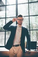 Cheerful young businessman. Confident young man in smart casual wear adjusting his eyeglasses and looking at camera with smile while standing in front of the big window in office or cafe photo
